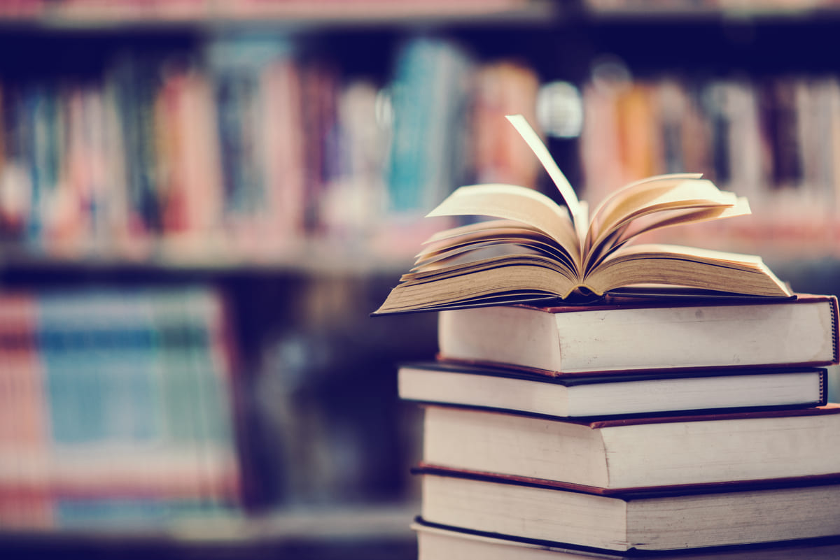 Otwarta książka na stosie tomów, w tle regały biblioteczne